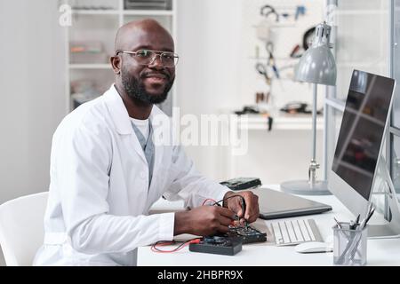 Junge fröhliche Werkstatt oder Laborarbeiter in Whitecoat Löten Details der Leiterplatte nach Arbeitsplatz und Blick auf Sie Stockfoto