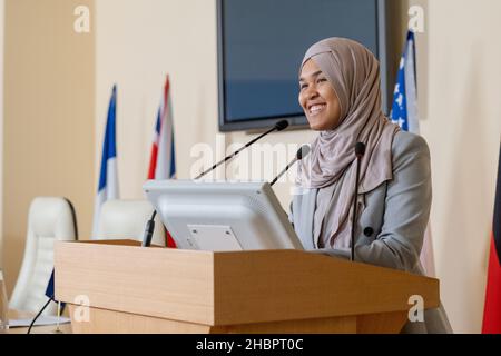 Junge erfolgreiche muslimische Geschäftsfrau im Hijab, die neben der Tribüne steht, während sie auf der Konferenz für ausländische Kollegen spricht Stockfoto