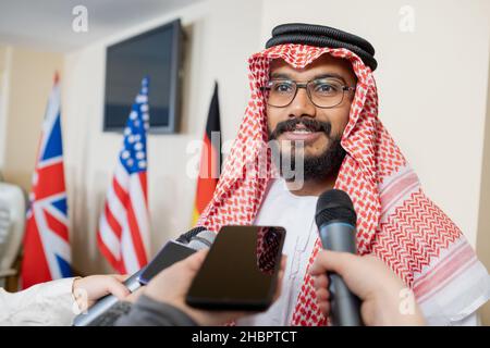 Junger selbstbewusster Delegierter in Nationalkleidung, der während der Pressekonferenz in einem Mikrofon von einem Journalisten gehalten wird Stockfoto