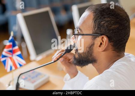 Junger, selbstbewusster Geschäftsmann oder Politiker, der im Konferenzsaal vor dem Publikum sitzt und mit einem Mikrofon spricht Stockfoto