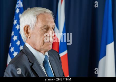 Gealterter grauhaariger Delegierter in formalwear, der vor der Kamera gegen Flaggen aus drei Ländern steht, die G7 aufgenommen wurden Stockfoto
