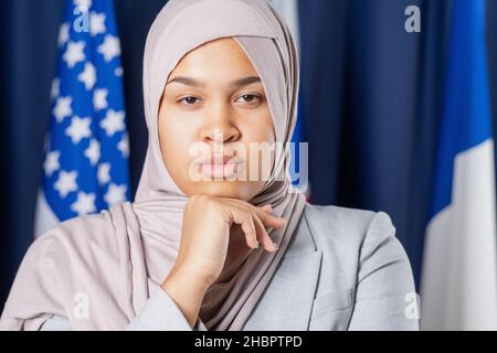 Junge ernsthafte weibliche Delegierte oder politische Führerin in Hijab, die die Kamera anschaut und dabei die Hand unter dem Kinn hält Stockfoto