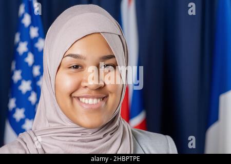 Junge, zeitgenössische muslimische Delegierte in Hijab, die mit einem Lächeln auf die Kamera blickt, während sie gegen Flaggen steht Stockfoto