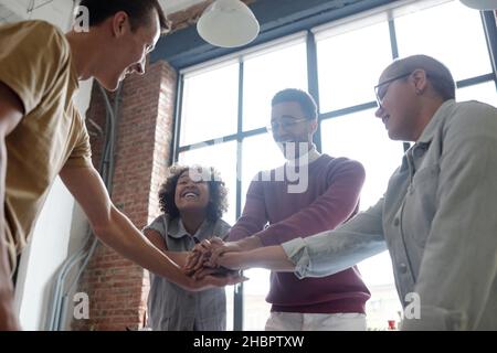 Fröhliche, moderne Mitarbeiter, die in einem großen Büro vor der Kamera stehen und einen Haufen Hände machen Stockfoto