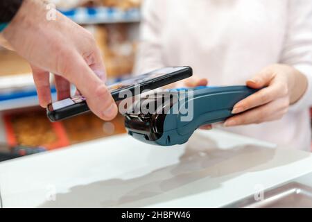 Die Hände einer Frau werden von einem Zahlungsterminal gehalten, und ein Mann zahlt für einen Kauf mit einem Smartphone. Seitenansicht. Das Konzept von NFC, Business und Banking t Stockfoto