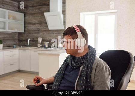 Ernsthafter Mann mit Kopfhörern, der Musik hört, während er sich im Rollstuhl im geräumigen Wohnzimmer oder in der Küche bewegt Stockfoto