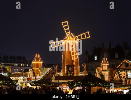 Deutscher Weihnachtsmarkt in der Nacht wunderschön beleuchtet hölzernen Straßenstände. Riesige beleuchtete Windmühle Käufer genießen ein festliches Weihnachtsgetränk Bier Stockfoto