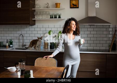 Lächelnde Frau mit Kaffeetasse, die in der Küche zu Hause wegschaut Stockfoto