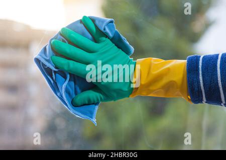 Eine Hand mit einem gelben und grünen Gummihandschuh wischt das schmutzige Glas eines Fensters mit einem nassen blauen Tuch ab. Im Hintergrund können Gebäude und Bäume sein Stockfoto