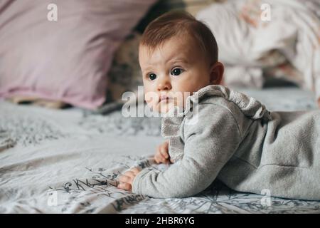 Seitenansicht eines kleinen Mädchens liegt auf ihrem Bauch auf dem Bett und schaut auf die Kamera. Neugeborenes Kind. Schönes Mädchen. Gesunder Lebensstil. Stockfoto