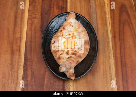 Traditionelle georgische Küche. Ajara khachapuri mit Ei auf schwarzem Teller auf Holzhintergrund Draufsicht, Nahaufnahme mit Kopierraum. Hochwertige Fotos Stockfoto