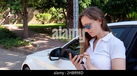 Eine Frau in einem weißen Kleid in der Nähe eines teuren Autos schreibt eine SMS an das Telefon ruft den Kundendienst oder schreibt an Freunde im sozialen Netzwerk. Stockfoto