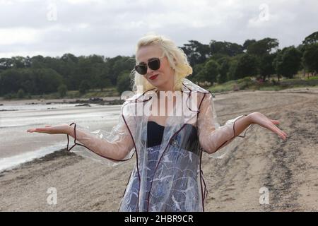 Ziemlich glamouröse junge Frau in einem Retro 1950s Badeanzug unter einem durchscheinenden Regenmantel aus Kunststoff mit lila Paspeln, lächelnd und einem emotionalen, leuchtend roten Lippenstift Stockfoto