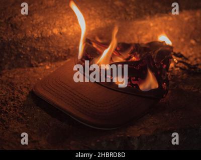 Eine dunkle Textilkappe brennt mit hellem Feuer, während sie nachts auf einer Betonoberfläche liegt. Bild in Brauntönen Stockfoto
