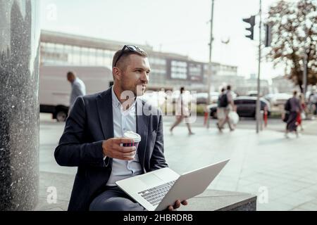 Ein junger Geschäftsmann hält seinen Laptop, während er Kaffee trinkt. Remote-Arbeit im Freien. Stockfoto
