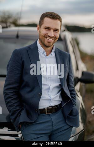 Junger lächelnder Mann mit schwarzem Oldtimer in schwarzem Anzug. Der Mensch steht in der Nähe seines Autos im Freien. Stockfoto