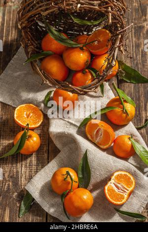 Mandarinen, die aus dem Korb fallen. Orangen, Mandarinen, Clementinen, Zitrusfrüchte) mit grünen Blättern. Holzhintergrund, rustikaler Stil, Draufsicht Stockfoto