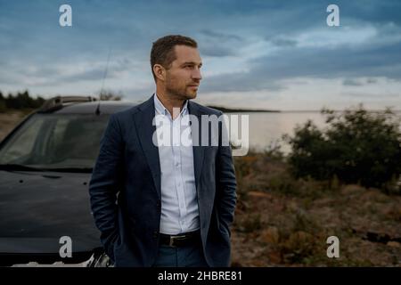 Junger, hübscher Mann mit schwarzem Oldtimer und schwarzem Anzug. Der Mensch steht in der Nähe seines Autos im Freien. Stockfoto