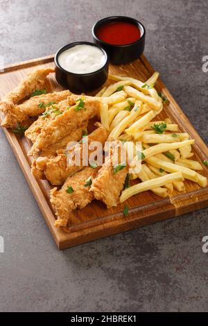 Hausgemachte knusprige Chicken Strips und Pommes Frites werden auf dem Holztablett auf einem Betontisch in der Nähe angezeigt. Vertikal Stockfoto
