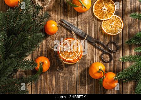 Trockene Orangen Scheiben Snack. Getrocknete Zitrusfrüchte für Weihnachtsdekorationen. Mandarinen mit Blättern, Tannenzweigen, Schere und Faden. Holzrückenboden Stockfoto