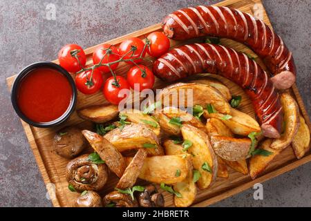 Gegrillte Würstchen im deutschen Stil mit Kartoffelkeilen und Pilzen und Sauce aus nächster Nähe auf einem Holztablett auf dem Tisch. Horizontale Draufsicht von oben Stockfoto