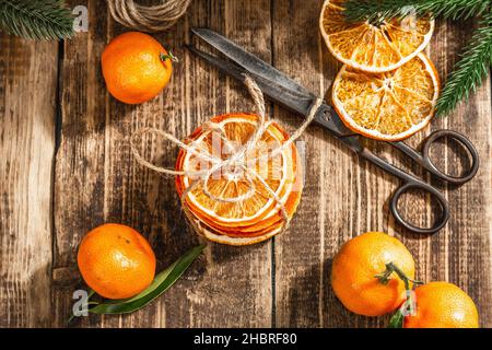 Trockene Orangen Scheiben Snack. Getrocknete Zitrusfrüchte für Weihnachtsdekorationen. Mandarinen mit Blättern, Tannenzweigen, Schere und Faden. Holzrückenboden Stockfoto