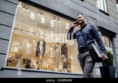 Junger Geschäftsmann, der seinen Laptop in der Hand hält, während er telefoniert. Gehen auf der Straße. Stockfoto