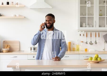 Ein glücklicher junger afroamerikanischer bärtiger Mann mit einem Glas sauberem Wasser spricht telefonisch Stockfoto