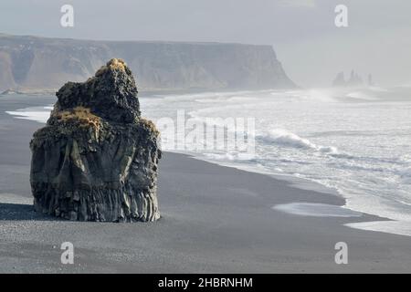 An einem dunklen vulkanischen Sandstrand in der Nähe von Dyrhólaey steht ein felsiger Überrest, der von mächtigen Wellen umspült wird und in Richtung entfernter Felsstapel bei Vik blickt Stockfoto