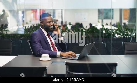 Fröhlicher schwarzer Geschäftsmann, der am Laptop arbeitet und ein Telefongespräch hat Stockfoto