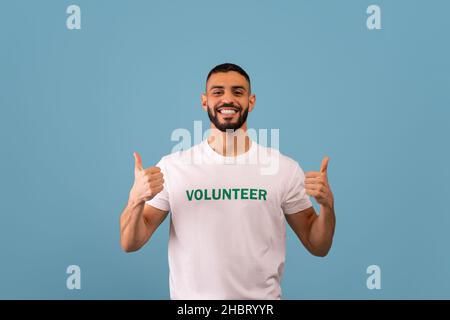 Freiwilligenarbeit. Glücklicher arabischer Aktivist im Freiwilligen-T-Shirt mit Daumen nach oben, lächelnd vor der Kamera, blauer Hintergrund Stockfoto