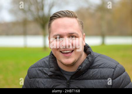 Lachender, fröhlicher junger Mann in einem Herbstpark in einem Kopf- und Schulterporträt, während er auf die Kamera schaut Stockfoto