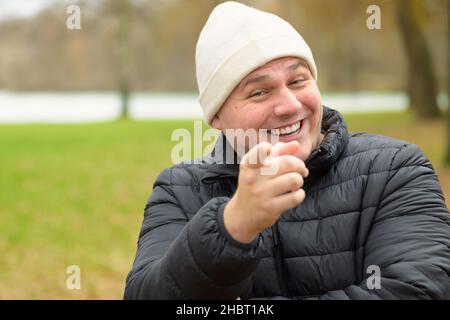 Lachender junger Mann in warmer Winterjacke und Mütze, der mit dem Finger auf die Kamera im Freien in einem Park zeigt Stockfoto