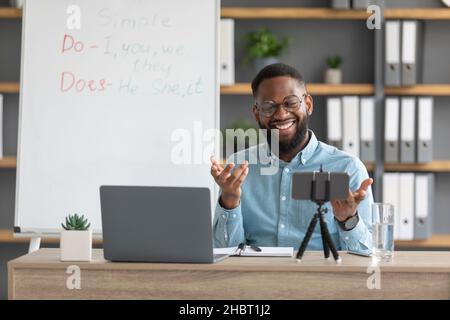 Frohes junges schwarzes männliches Lehramt, Tutor in der Brille schaut Laptop und Handy Webcam im Home Office Interieur mit Tafel Stockfoto