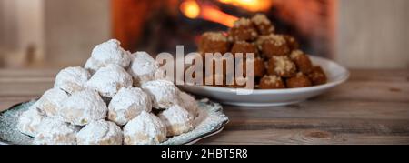 Griechische Weihnachtsdesserts. Traditionelle hausgemachte Kourabiedes und melomakarona auf einem Tisch, brennenden Kamin Hintergrund Stockfoto