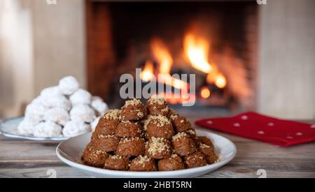 Griechische Weihnachtsdesserts. Traditionelle hausgemachte melomakarona und kourabiedes auf einem Tisch, brennenden Kamin Hintergrund Stockfoto