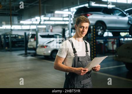 Mittellange Aufnahme eines lächelnden, gut aussehenden jungen Mechanikers mit uniform getragenen Clipboard, der in der Werkstatt der Autowerkstatt steht. Stockfoto