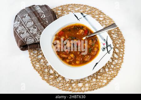 Reichhaltige und würzige ländliche rote Suppe mit Schweinefleisch, rotem Pfeffer, Tomaten, Pilzen, Gemüse und Bohnen auf dem Teller, Handtuch und Bambuskissen auf weißem Hintergrund. Stockfoto