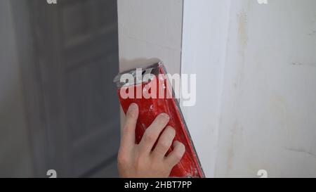 Die Wand mit einer Maschine und Schleifpapier schleifen. Bulgarisch. Gipser glättet die Wände mit Sandpapier aus nächster Nähe. Männliche Hand eines Arbeiters.Hausrenovierung c Stockfoto
