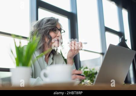Reife Geschäftsfrau, die Sprachnachrichten auf dem Mobiltelefon in der Nähe des Laptops aufzeichnete und verschwommene Kaffeetassen im Büro aufzeichnete Stockfoto
