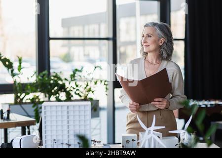 Eine reife Geschäftsfrau hält einen Papierordner in der Nähe des Headsets und von Modellen von Gebäuden im Büro Stockfoto