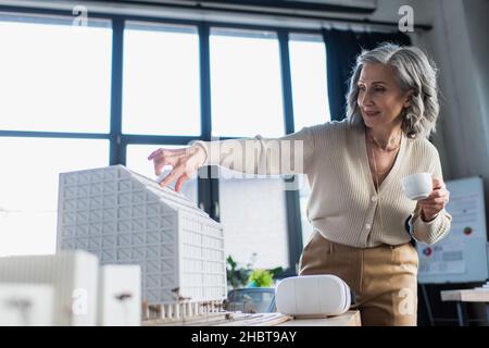 Eine reife Geschäftsfrau hält eine Tasse Kaffee in der Nähe von Modellen von Gebäuden und einem vr-Headset im Büro Stockfoto