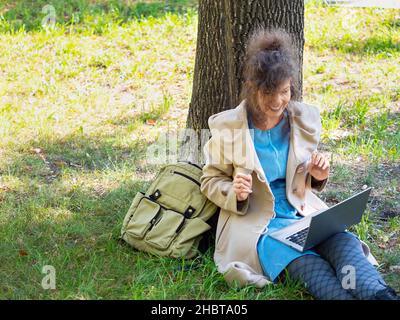 Foto einer Frau mittleren Alters in Kopfhörern mit Laptop im Stadtpark während eines Online-Videoanrufs. Online-Arbeiten oder E-Learning-Konzept. Stockfoto