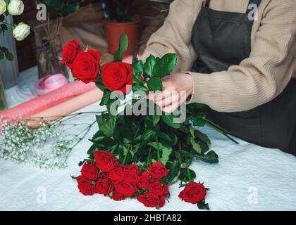 Frau Floristin Schaffung eines Straußes von frischen roten Rosen. Zugeschnittenes Bild einer weiblichen Floristin, die Rosenblumen mit Werkzeugen anordnet. Konzept der Arbeit mit Blumen, Blumengeschäft. Selektiver Fokus Stockfoto