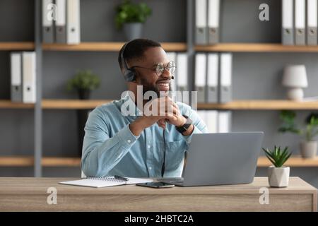 Zufriedener, tausendjähriger, schwarzer bärtiger Mann in Brille und Kopfhörer arbeitet am Laptop und schaut auf leeren Raum Stockfoto