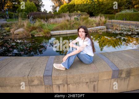 Teenager-Frau, die ihr Smartphone an einem mit Lilly gefüllten Teich benutzt Stockfoto