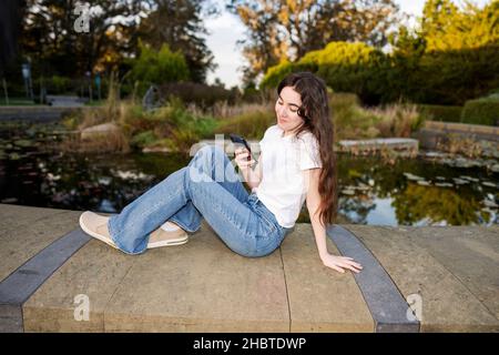 Teenager-Frau, die ihr Smartphone an einem mit Lilly gefüllten Teich benutzt Stockfoto