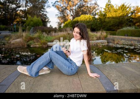 Teenager-Frau, die ihr Smartphone an einem mit Lilly gefüllten Teich benutzt Stockfoto