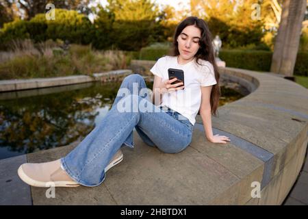 Teenager-Frau, die ihr Smartphone an einem mit Lilly gefüllten Teich benutzt Stockfoto