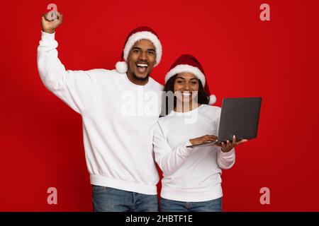 Emotionale schwarze Liebhaber in Santa Hüte halten Laptop Stockfoto